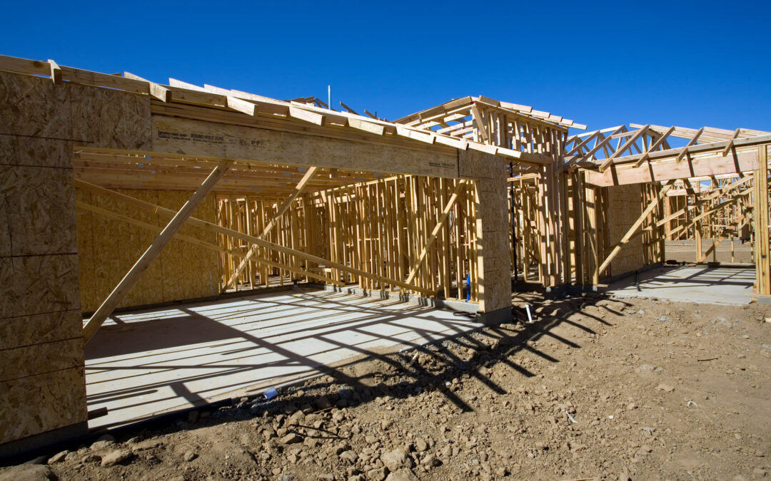 A single-story wood-framed house stands on a fresh concrete slab at a sunny construction site under a clear blue sky.